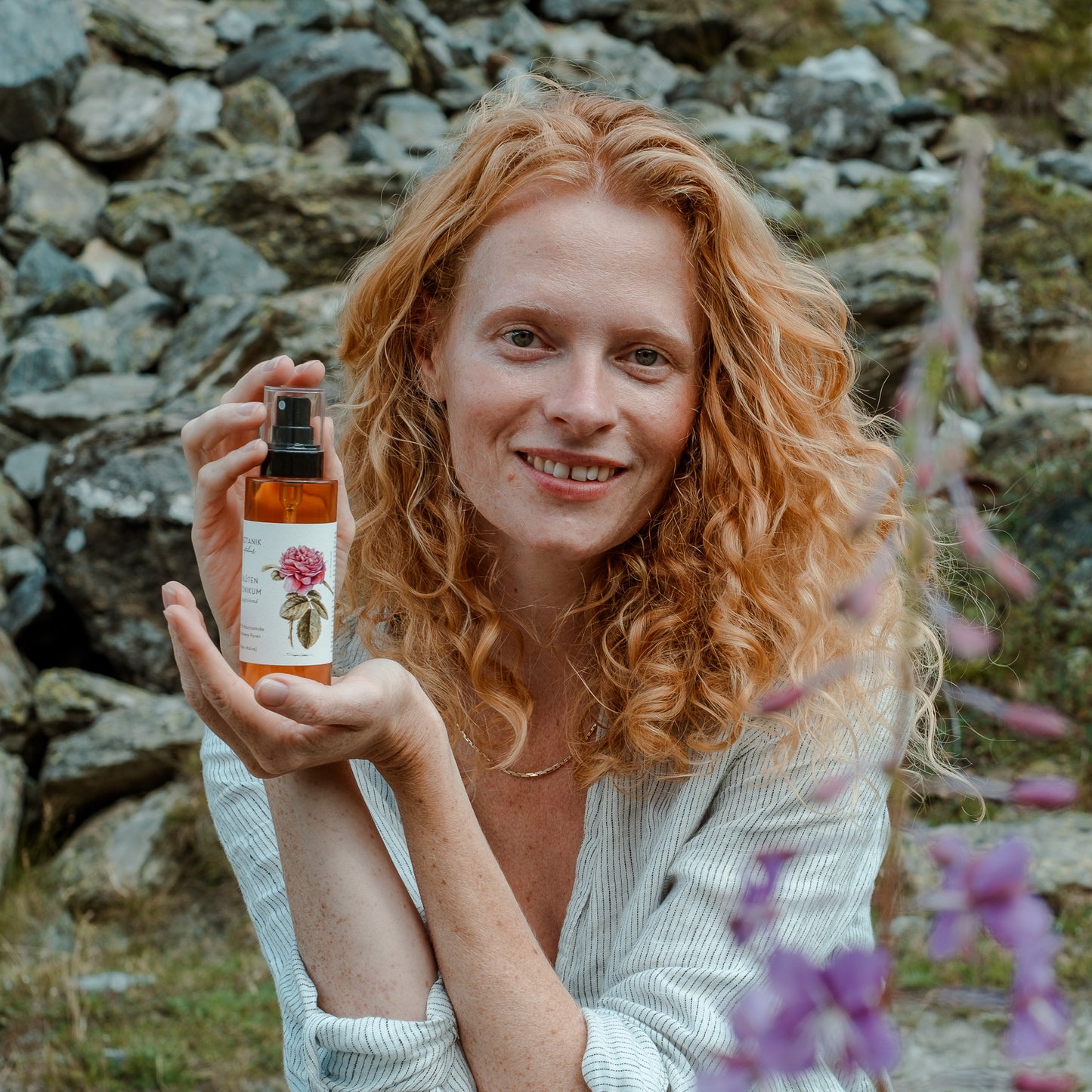 Frau mit roten Locken blickt in die Kamera, lächelt, und zeigt das Blüten Tonikum mit 5% Niacinamide von Botanik Atelier.