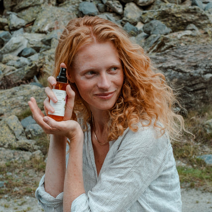 Eine Frau mit roten locken hält das Blüten Tonikum mit 5% Niacinamide von Botanik Atelier neben ihr lächelndes Gesicht.